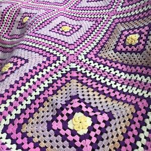 May include: A handmade crochet blanket in shades of purple, lavender, and white. The blanket features a diamond pattern with yellow flower accents. The texture is visible, showcasing the intricate stitch work. The overall design is a cozy, handcrafted item.
