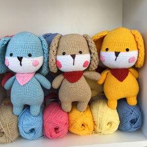 Op de afbeelding: Drie gehaakte konijnenpoppen in blauw, beige en geel, elk met slappe oren, een witte snuit en roze wangen. Ze dragen bandana's en staan boven kleurrijke bollen wol. De poppen zijn handgemaakt en geschikt voor kinderen.