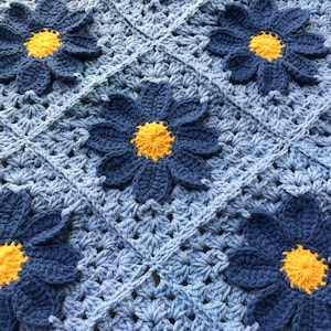 May include: A crocheted baby blanket with a blue and yellow color scheme. The blanket features a repeating pattern of blue flowers with yellow centers.