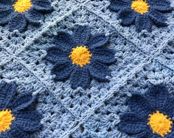 Op de afbeelding: Een gehaakt babydekentje in blauw en geel. Het dekentje heeft een herhalend patroon van blauwe bloemen met gele centra.