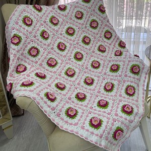 May include: A white crocheted blanket with a pink and green floral pattern. The blanket is draped over a chair.