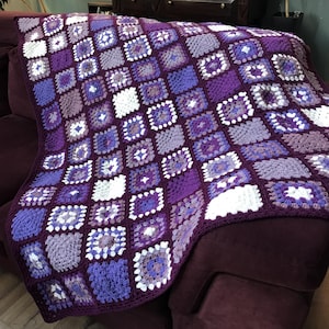Op de afbeelding: Een handgemaakte gehaakte deken in tinten paars, lavendel en wit. De deken heeft een vierkant patroonontwerp, waarbij elke vierkant een andere variant van het kleurenpalet laat zien. De deken is over een bordeauxrode bank gedrapeerd.