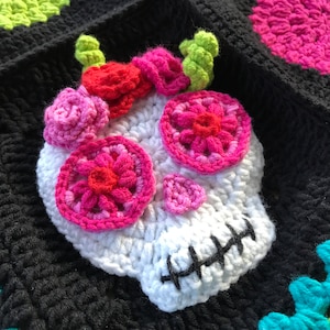 May include: White crocheted skull with pink and red flower embellishments. The skull has a black stitched mouth and is set against a black background.