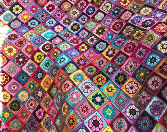 Op de afbeelding: Een kleurrijk gehaakt deken met een vierkant patroon van bloemen in verschillende kleuren, waaronder roze, paars, blauw, groen en geel.