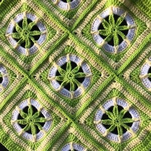 May include: A handmade crochet blanket featuring a repeating pattern of green, white, and cream-colored squares. Each square has a central floral design in green, surrounded by a white circle and cream-colored border. The blanket is made of soft yarn.