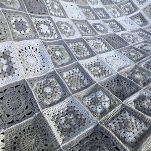 Op de afbeelding: Een handgemaakte gehaakte deken met een patchwork-ontwerp van vierkante motieven in grijstinten en wit. De deken toont ingewikkelde bloemen- en geometrische patronen, waardoor een textuur en gezellige uitstraling ontstaat. Het algehele ontwerp is een mix van licht- en donkergrijze vierkanten.