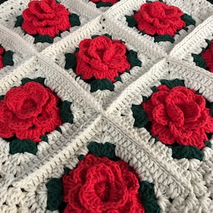 May include: A crocheted blanket with a repeating pattern of red flowers and green leaves on a cream background.
