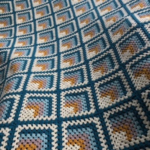May include: A crocheted blanket with a repeating pattern of squares in shades of blue, white, beige, and gold.
