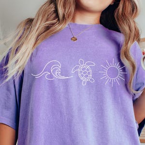 May include: A light purple t-shirt featuring a white graphic of a wave, a turtle, and a sun. The shirt is a relaxed fit. A person is holding a mug with a sun design.