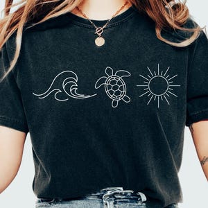 May include: Black t-shirt featuring a white graphic of a wave, a sea turtle, and a sun. The shirt is made of a soft material and has a relaxed fit. The design is simple and evokes a beach or ocean theme.