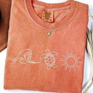 May include: Coral-colored Comfort Colors t-shirt with a white graphic of a wave, a turtle, and a sun. A gold necklace and a brown hat are next to the shirt. The shirt tag reads "Comfort Colors".