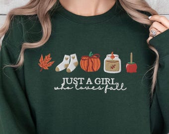 Könnte beinhalten: Dunkelgrünes Sweatshirt mit gestickten Herbstsymbolen: ein Blatt, Socken, Kürbis, Glas und Apfel. Der Text "JUST A GIRL who loves fall" ist in weißer Schrift geschrieben. Das Sweatshirt hat einen Rundhalsausschnitt und lange Ärmel.