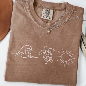 May include: Brown Comfort Colors t-shirt with a white wave, turtle, and sun graphic. The shirt has a crew neck and short sleeves. A silver necklace with a pendant is draped across the shirt.