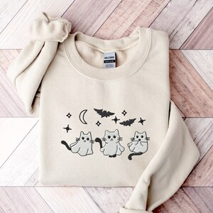May include: A cream-coloured sweatshirt with a black graphic of three cats dressed as ghosts with bats and stars in the background.