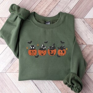 May include: A green sweatshirt with an embroidered Halloween design. The design features four orange jack-o'-lanterns, each with a black cat inside. Black bats and stars are also embroidered around the pumpkins. The sweatshirt has long sleeves.