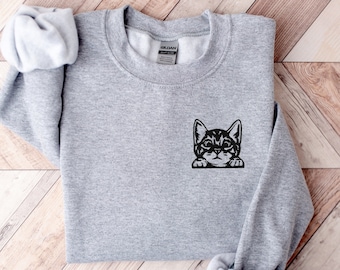 Op de afbeelding: Een grijs sweatshirt met een zwart geborduurd beeld van een kat die over de rand van de zak kijkt.