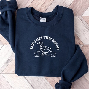Può includere: Una felpa blu navy con una grafica bianca di un'anatra e due anatroccoli con il testo "Let's get this bread".