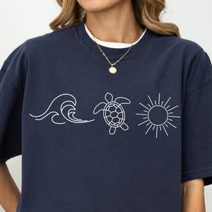 May include: Navy blue t-shirt with a white graphic of a wave, a turtle, and a sun. The shirt has short sleeves and a relaxed fit. The design is simple and evokes a beach or ocean theme.