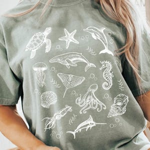 May include: Sage green t-shirt featuring a white illustration of various sea creatures, including a turtle, starfish, dolphin, and octopus. The shirt is a relaxed fit and has short sleeves. The design is a detailed line drawing of marine life.