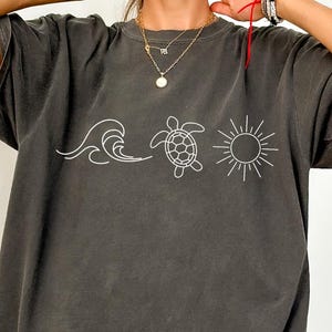 May include: Dark gray t-shirt featuring a white outline design of a wave, a turtle, and a sun. The shirt has short sleeves and a relaxed fit. The design is centered on the front of the shirt, creating a casual, beach-themed aesthetic.