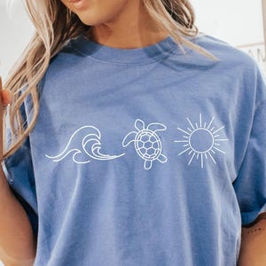May include: A light blue t-shirt with a white graphic of a wave, a turtle, and a sun. The person is wearing white socks with black stripes and holding a glass of iced coffee. The shirt is a relaxed fit.