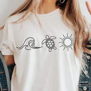 May include: Cream-colored t-shirt featuring a black line drawing of a wave, a turtle, and a sun. The shirt has short sleeves and a relaxed fit. The model is wearing a denim jacket.