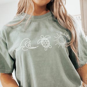 May include: Sage green t-shirt featuring a white graphic of a wave, a turtle, and a sun. The shirt is a relaxed fit and has short sleeves. The person is wearing white shorts and white socks with black stripes.