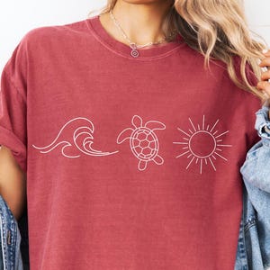 May include: A maroon t-shirt featuring a white line drawing of a wave, a turtle, and a sun. The shirt has short sleeves and a crew neck. A denim jacket is draped over the shoulders.