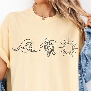 May include: A light yellow t-shirt featuring a black line drawing of a wave, a sea turtle, and a sun. The shirt has short sleeves and a relaxed fit. The model is wearing a denim jacket.
