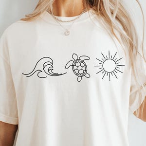 May include: Cream-colored t-shirt featuring a black line art design of a wave, a sea turtle, and a sun. The shirt has short sleeves and a relaxed fit, suitable for casual wear. The design evokes a beach or ocean theme.