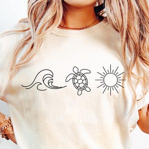 May include: Cream-colored t-shirt featuring a black line drawing of a wave, a turtle, and a sun. The shirt is made of soft material and is paired with blue jeans. The design evokes a beach or ocean theme.