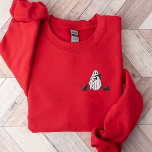 May include: Red sweatshirt with a white ghost holding a cat and two black cats on the front. The sweatshirt has a crew neck and long sleeves.