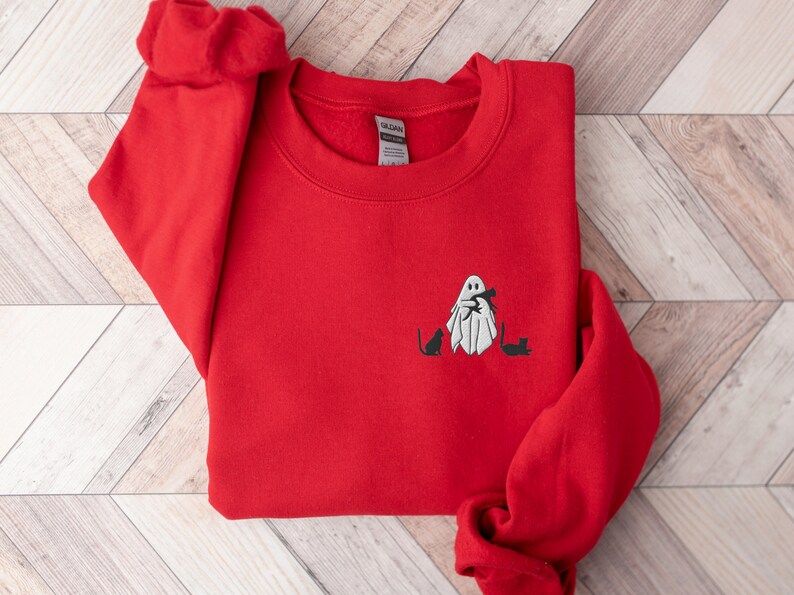 May include: Red sweatshirt with a white ghost holding a cat and two black cats on the front. The sweatshirt has a crew neck and long sleeves.
