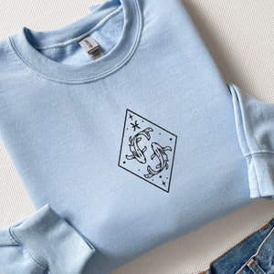 Puede incluir: Una sudadera azul claro con un contorno negro del s&iacute;mbolo del zod&iacute;aco de Piscis, dos peces y estrellas dentro de un diamante.