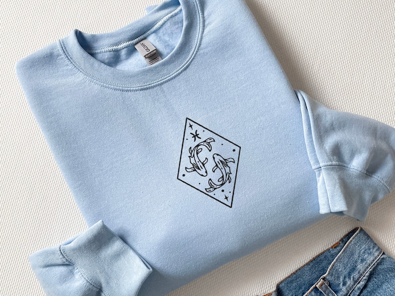 Puede incluir: Una sudadera azul claro con un contorno negro del s&iacute;mbolo del zod&iacute;aco de Piscis, dos peces y estrellas dentro de un diamante.