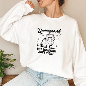 Op de afbeelding: Witte longsleeve trui met een zwarte afbeelding van een kat met een bril en de tekst "Undiagnosed but somethin ain't right".