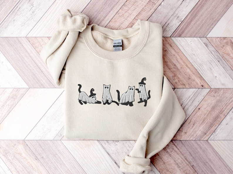 May include: A cream-colored sweatshirt with an embroidered design of five cats dressed as ghosts for Halloween.