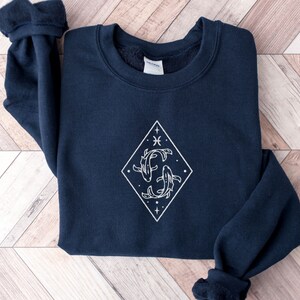 Puede incluir: Una sudadera azul marino con un gr&aacute;fico blanco de dos peces nadando dentro de un diamante. El gr&aacute;fico tambi&eacute;n incluye estrellas y el s&iacute;mbolo del zod&iacute;aco de Piscis.