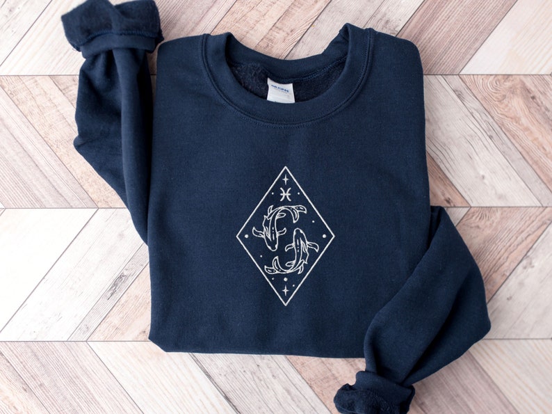 Puede incluir: Una sudadera azul marino con un gr&aacute;fico blanco de dos peces nadando dentro de un diamante. El gr&aacute;fico tambi&eacute;n incluye estrellas y el s&iacute;mbolo del zod&iacute;aco de Piscis.
