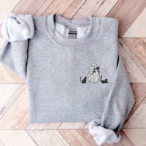 May include: A gray sweatshirt with a ghost holding a cat and two other cats sitting on the ground. The ghost and cats are black.