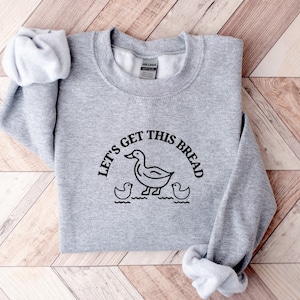 May include: Gray sweatshirt with a black graphic of a duck and the text "Let's get this bread".