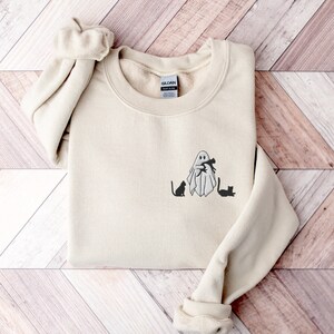 May include: A beige sweatshirt with a black ghost holding two black cats. The ghost is wearing a black hat and has a black mouth. The text "Gildan" is visible on the tag.