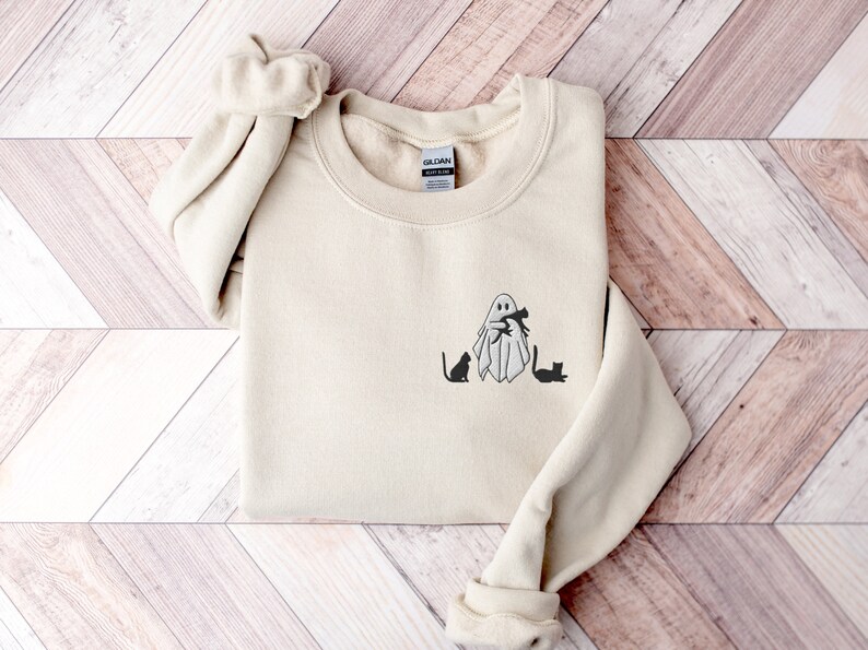 May include: A beige sweatshirt with a black ghost holding two black cats. The ghost is wearing a black hat and has a black mouth. The text "Gildan" is visible on the tag.
