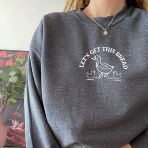 May include: A dark gray sweatshirt with a white graphic of ducks and the text "Let's Get This Bread".