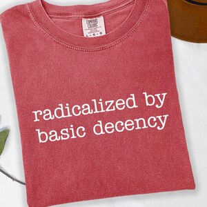 Puede incluir: Una camiseta roja con la frase blanca "radicalized by basic decency". La prenda tiene cuello redondo y una etiqueta que dice "Comfort Colors".