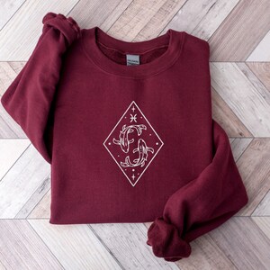Puede incluir: Una sudadera color burdeos con un gr&aacute;fico blanco de dos peces nadando dentro de un diamante. El gr&aacute;fico tambi&eacute;n incluye una estrella, una luna creciente y un s&iacute;mbolo que se parece al s&iacute;mbolo astrol&oacute;gico de Piscis.