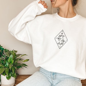 Puede incluir: Sudadera blanca con un gr&aacute;fico en blanco y negro de dos peces en forma de diamante. El gr&aacute;fico tambi&eacute;n incluye una estrella y un s&iacute;mbolo que parece un pez.