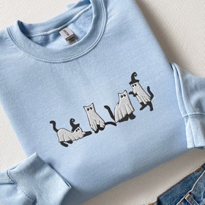 May include: A light blue sweatshirt with a white embroidered design of four cats dressed as ghosts for Halloween.
