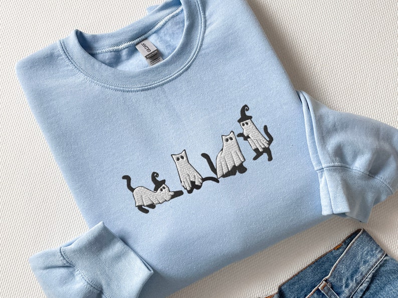 May include: A light blue sweatshirt with a white embroidered design of four cats dressed as ghosts for Halloween.