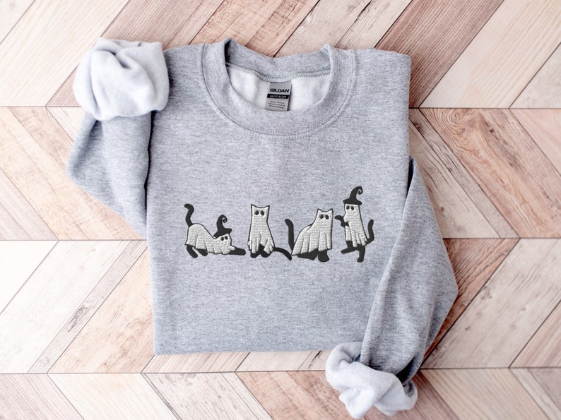 May include: A gray sweatshirt with a design of five cats dressed as ghosts for Halloween. The cats are in a line, with one cat wearing a witch's hat.